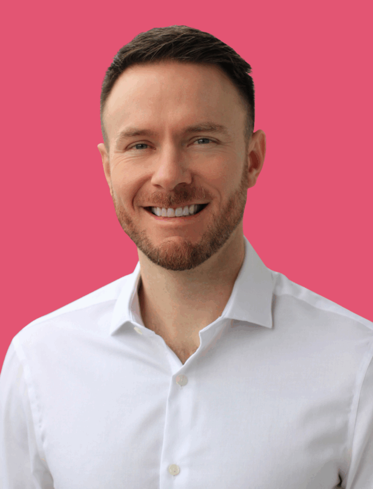 Smiling man with short brown hair and a trimmed beard wearing a white button-up shirt, against a solid pink background.